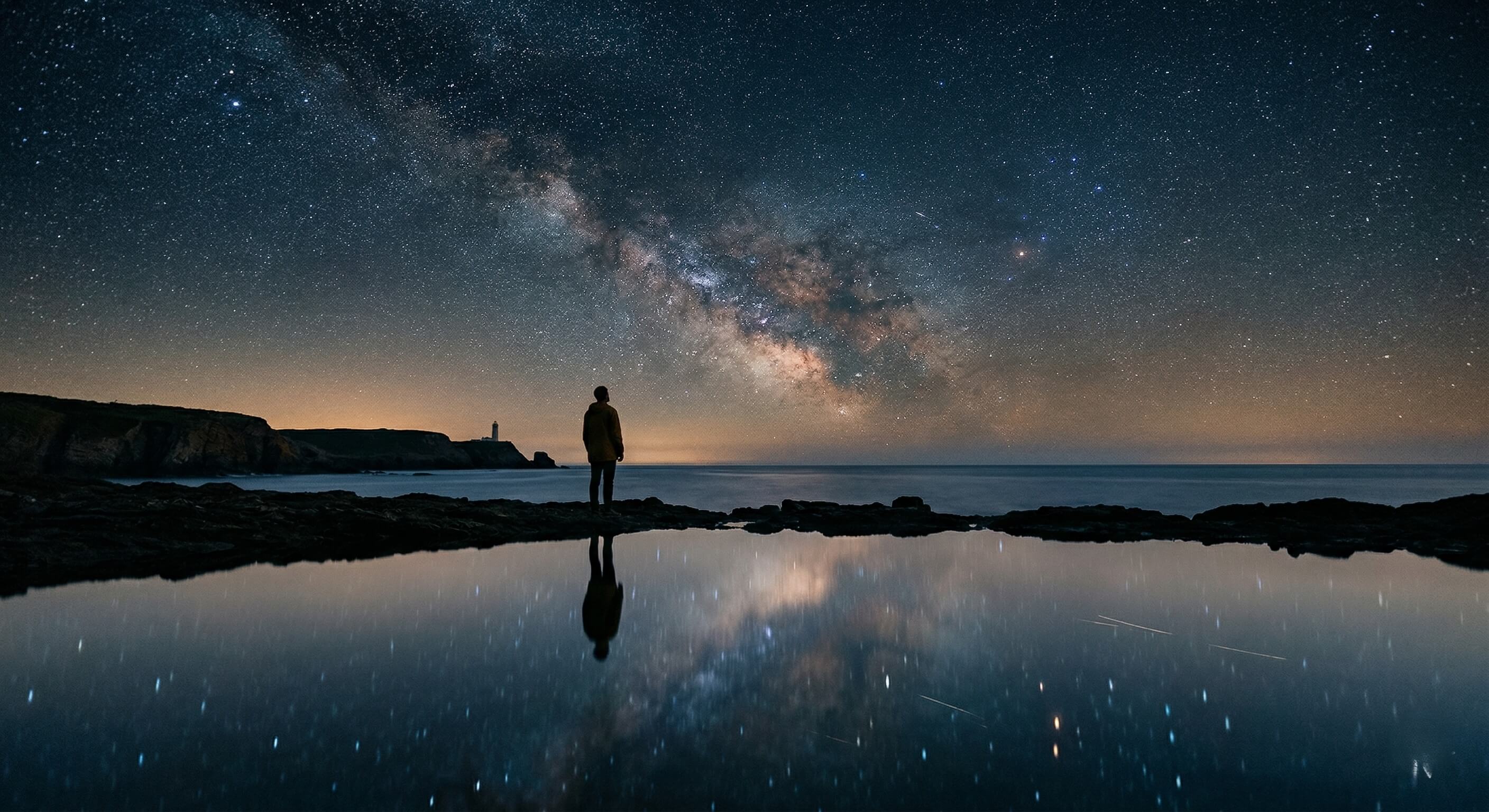 A person standing on a rocky formation by the sea, observing the Milky Way in the night sky reflected on the calm water, creating a mirror-like effect between sky and earth.